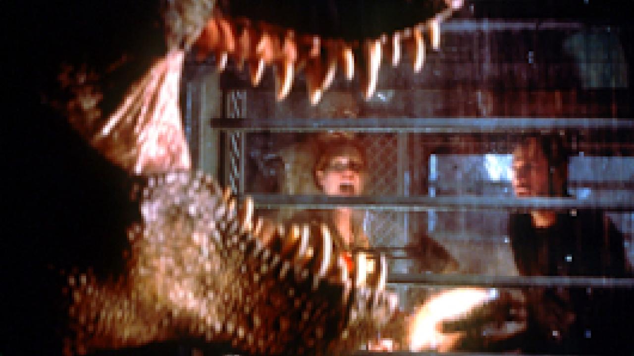 The Lost World: Jurassic Park, Szenenbild: Großaufnahme eines Tyranosaurus Rex mit geöffneten Maul, im Hintergrund sind zwei Menschen zu sehen.