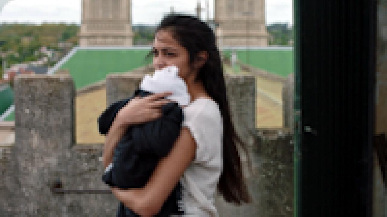 Maternal, Szenenbild: Eine junge Frau mit langen, schwarzen Haaren steht im Freien und hält ein Baby an sich gedrückt.