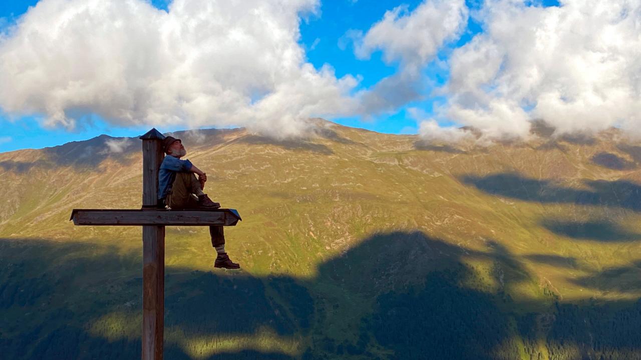 Märzengrund, Szenenbild: Ein Mann sitzt vor Alpenpanorama auf einem Gipfelkreuz.