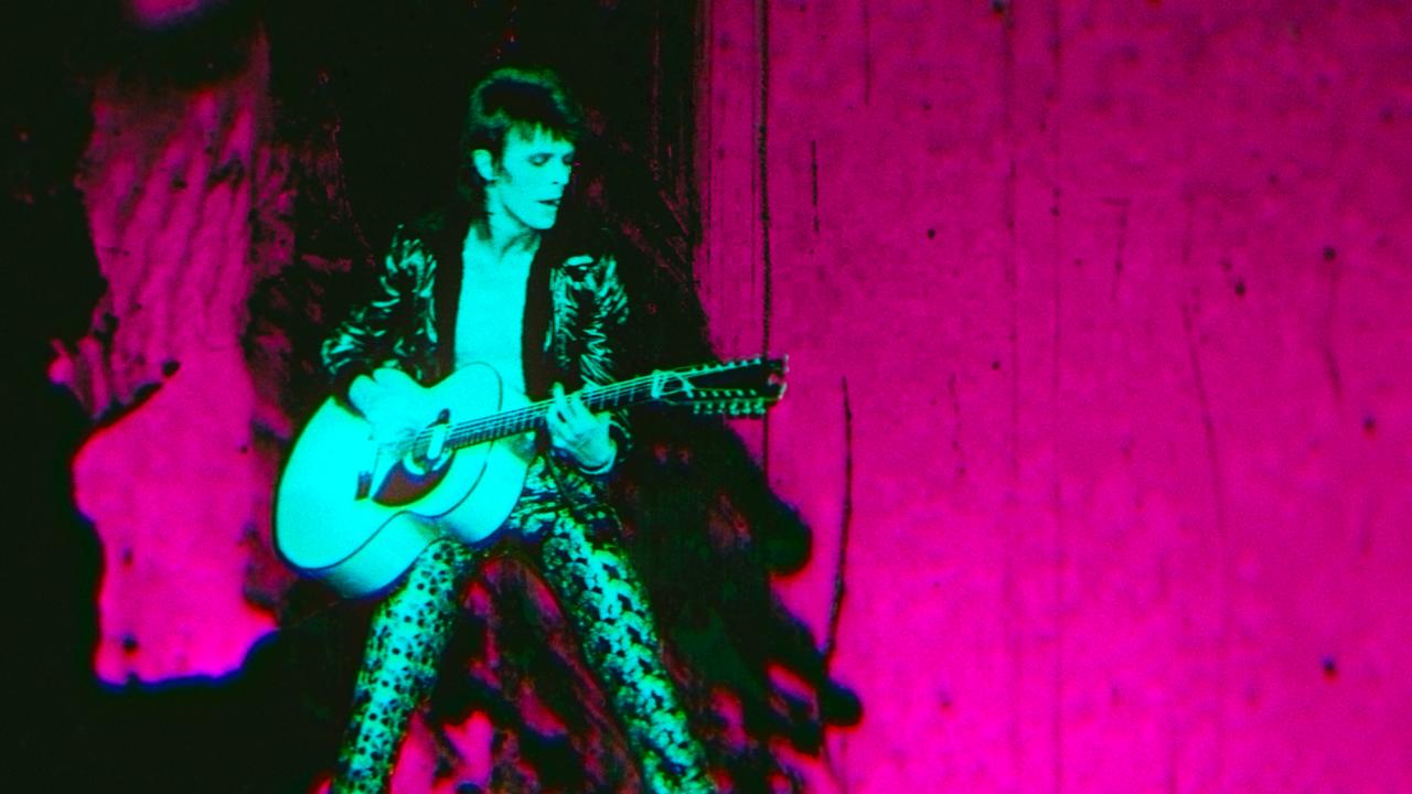 Moonage Daydream, Szenenbild: David Bowie in den 1970ern steht mit einer Gitarre auf einer Bühne. Er selbst ist ein einem grünlichen Ton, der Hintergrund in einem knalligen Pink gehalten.