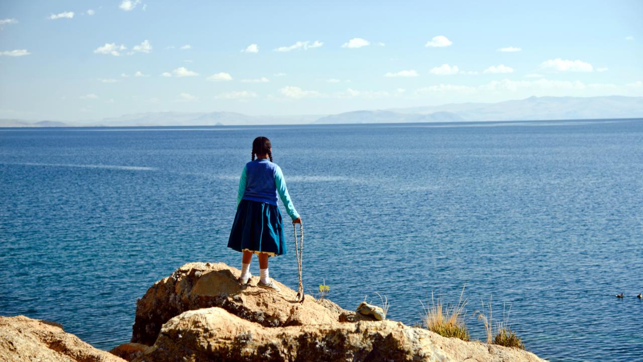 Die Tochter der Sonne, Szenenbild: Ein blau gekleidetes Mädchen steht mit dem Rücken zu den Betrachtenden auf einem Felsen. Vor ihr erstreckt sich der Titicacasee und die Weite des Himmels.