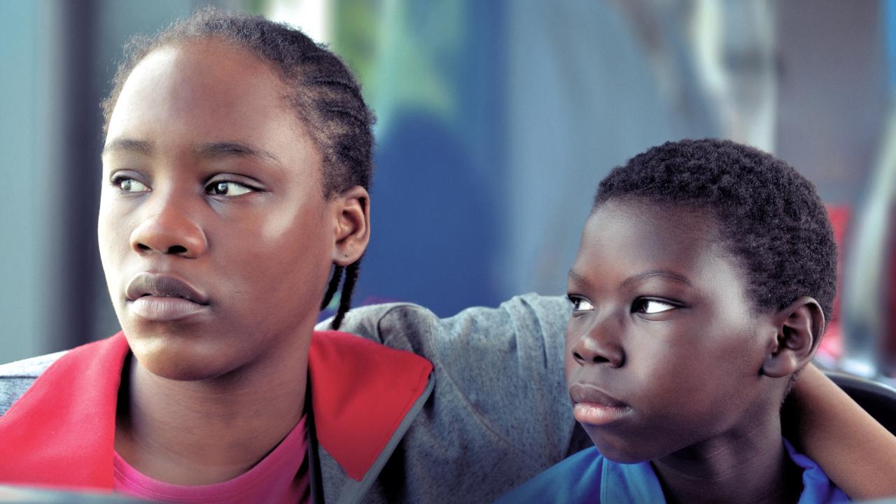 Tori & Lokita, Szenenbild: Nahaufnahme: Ein Mädchen im Teenageralter und ein jüngerer Junge, beide Personen of Color, sitzen nebeneinander. Das Mädchen hat ihren Arm um den Jungen gelegt.