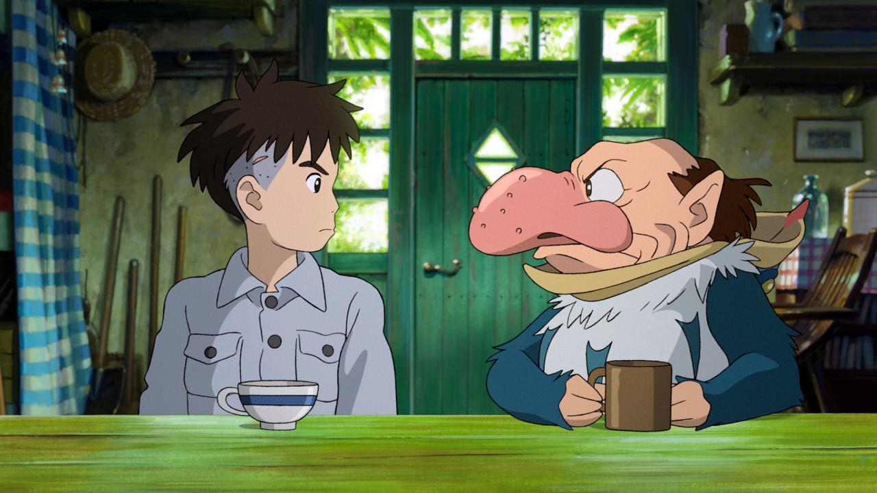 Der Junge und der Reiher, Szenenbild aus dem Anime: Ein Junge in grauem Hemd und ein Mann in einem Vogelkostüm sitzen an einem grünen Tisch und schauen sich grimmig an.