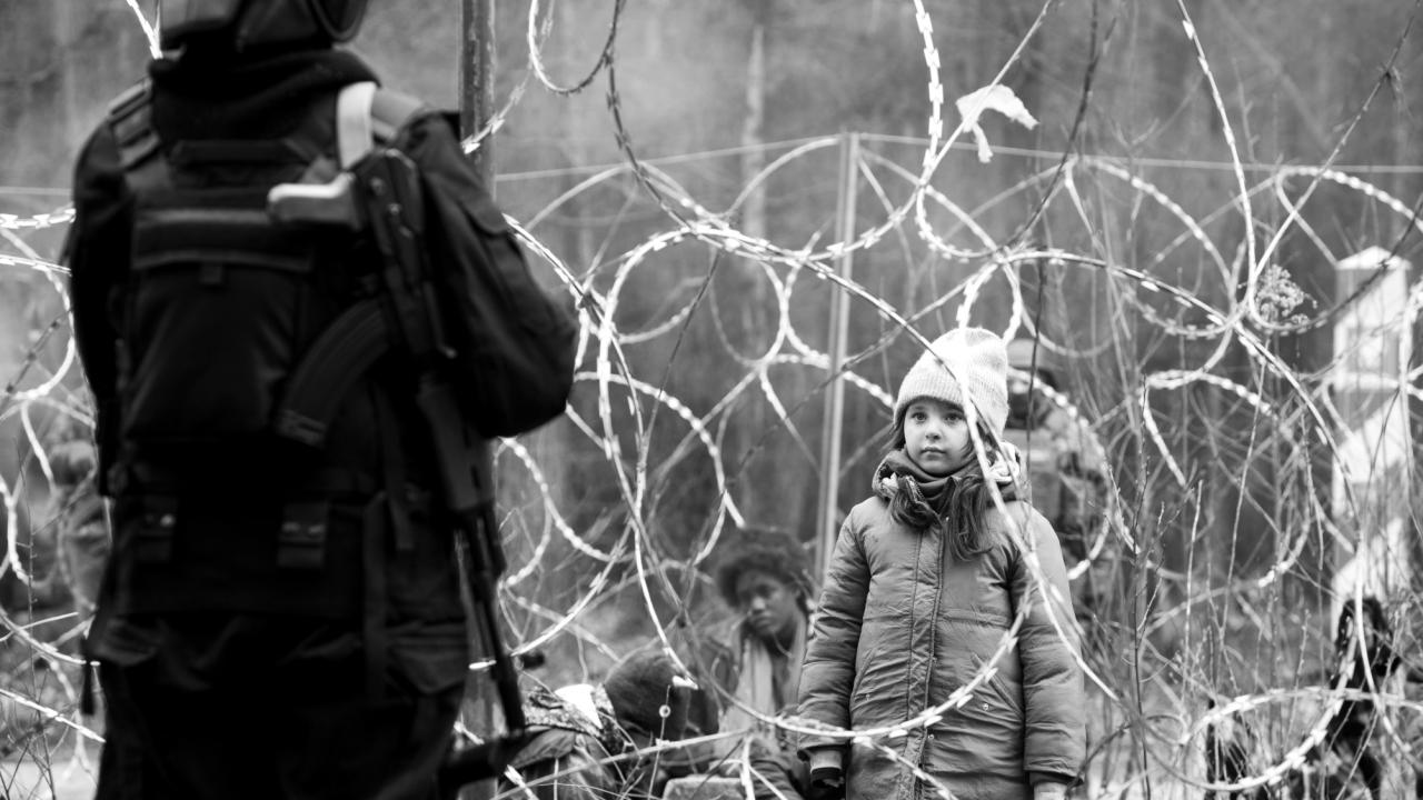 Green Border, Szenenbild: Ein junges Mädchen steht hinter einer Stacheldrahlabsperrung, im Vordergrund ist unscharf eine bewaffnete Person zu sehen, hinter dem Kind ebenfalls unscharf weitere Personen