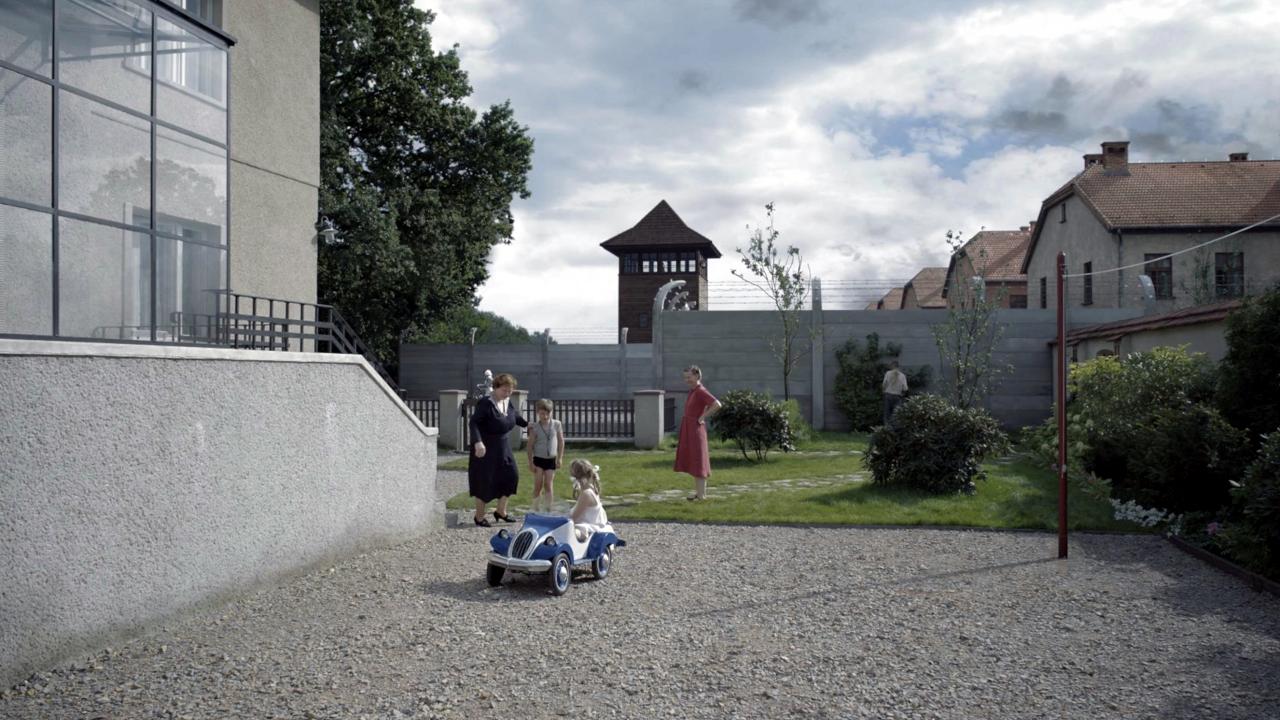 The Zone of Interest, Szenenbild: Zwei Frauen und zwei Kinder stehen neben einem Wohnhaus in einem Garten, der von einer hohen Mauer begrenzt ist. Im Hintergrund sind Gebäude und der Wachturm eines Konzentrationslagers zu sehen.