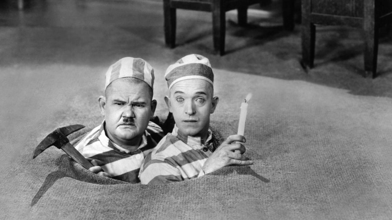 Szenenbild aus der Komödie Dick und Doof: Dem Henker entronnen: Die Komiker Oliver Hardy mit Schnauzbart und Stan Laurel schauen durch ein Loch im Boden eines Zimmers. Beide tragen gestreifte Sträflingsuniformen. Oliver Hardy häl