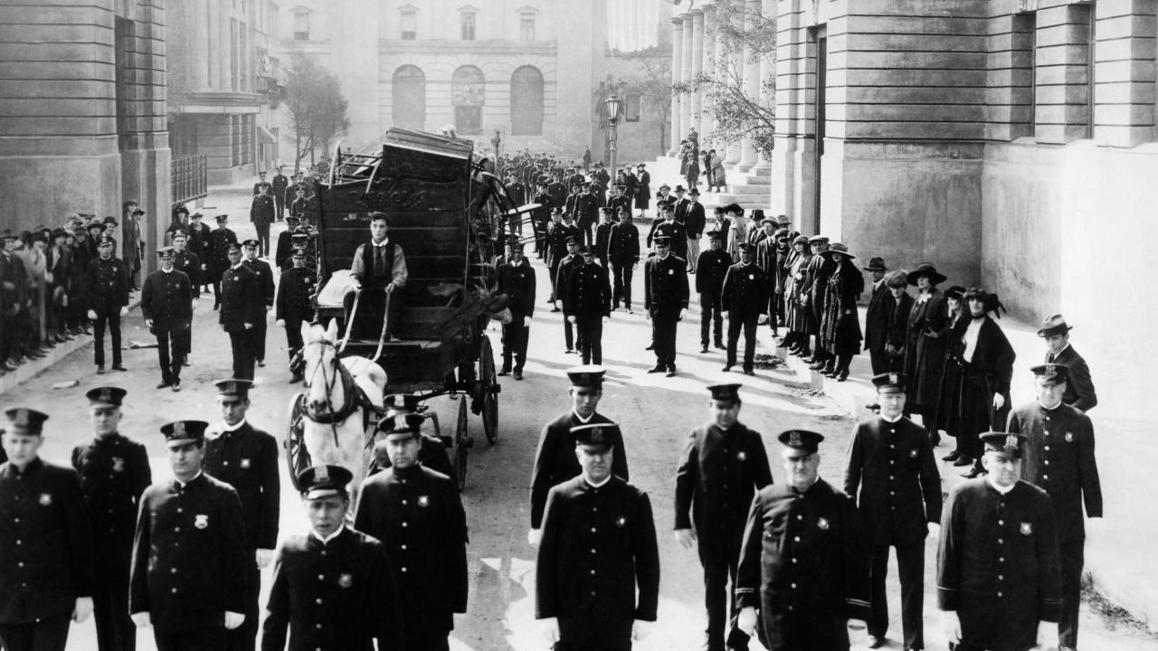 Schwarz-weißes Szenenbild aus dem Stummfilm Buster und die Polizei (USA 1922): Ein Straßenzug in Los Angeles mit unzählig vielen Polizisten in Uniform. In der Mitte des Bildes steht eine Pferdekutsche. Der Schauspieler Buster Kea