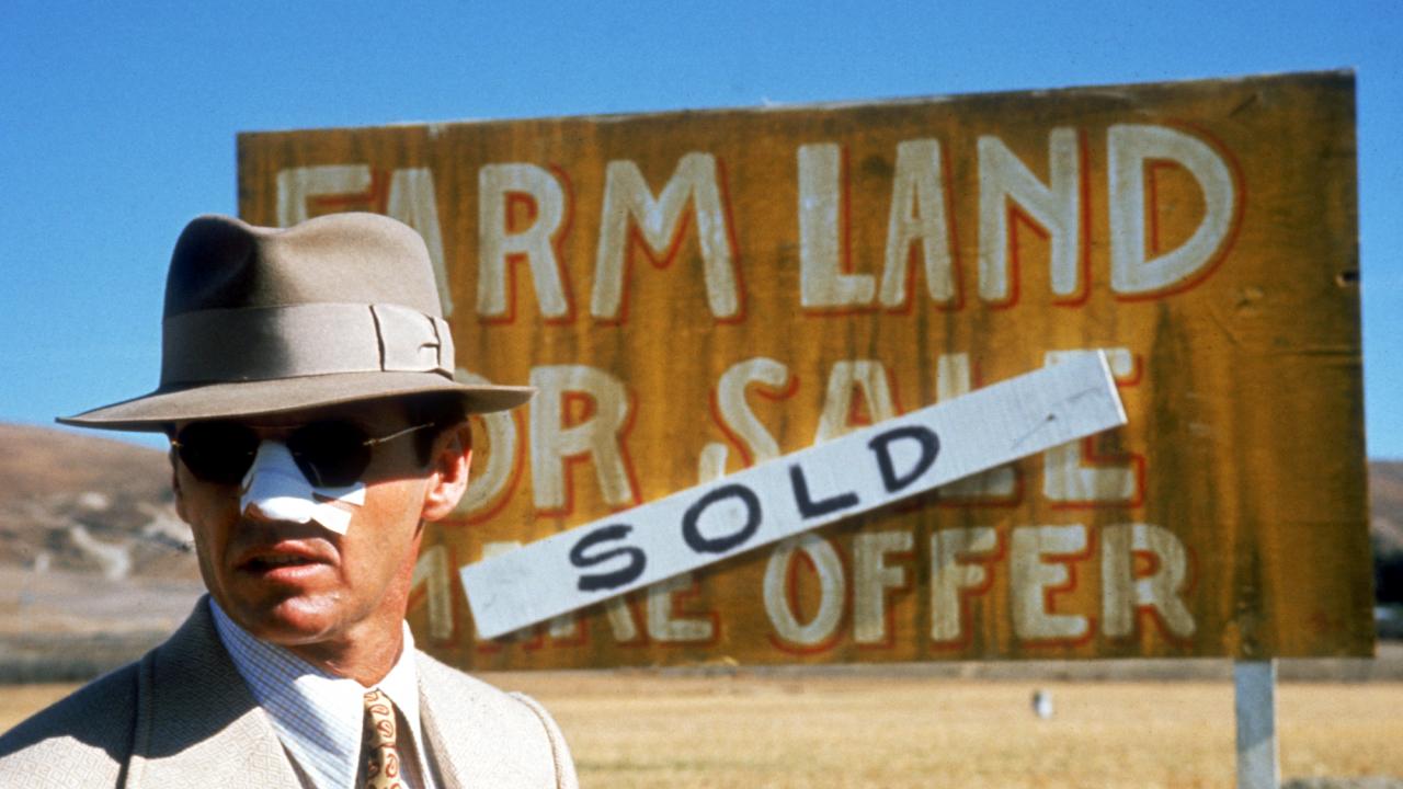 Szenenbild aus dem Neo-Noir-Film "Chinatown": Ein Mann, gekleidetim Stil der 1930er, steht vor einem Schild, auf dem "Farm Land for Sale" steht. Darüber ist ein Hinweis, dass das Land bereits verkauft wurde. Der Mann trägt ein Pflaster auf der Nase.