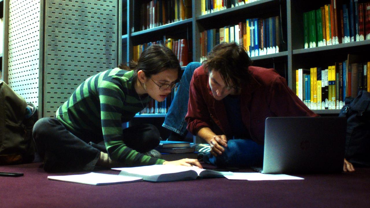 Szenenbild aus dem Drama "Die Gleichung ihres Lebens": Eine junge Frau und ein etwa gleichaltriger Mann sitzen nebeneinander auf dem Boden einer Bibliothek und arbeiten zusammen an Büchern und einem Laptop.