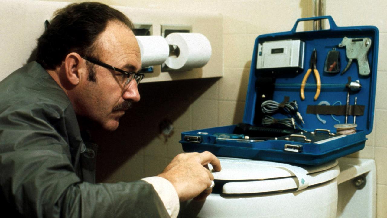 Szenenbild aus dem Thriller "Der Dialog": In einer weißgefließten Toilette hockt ein Mann mit hoher Stirn, Schnurrbart und Brille vor dem WC und schaut auf einen Elektronikwerkzeugkasten, der aufgeklappt vor ihm auf dem Klodeckel steht