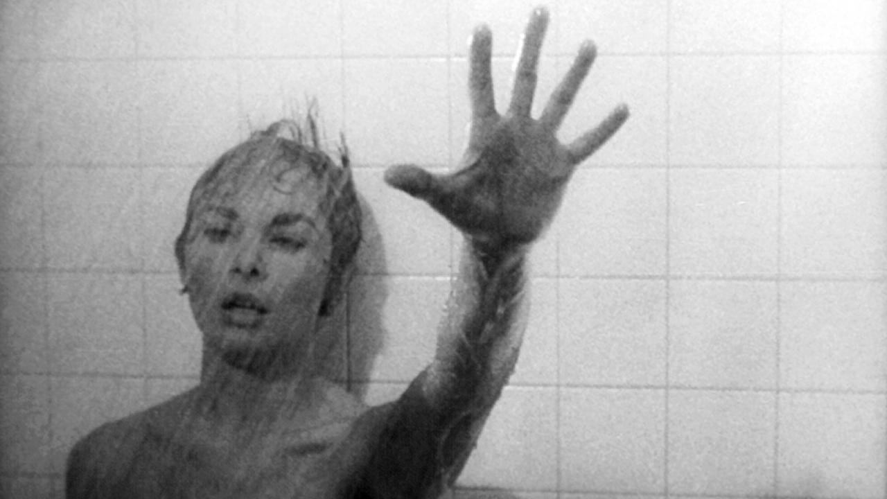 Szenenbild aus dem Film PSYCHO: Eine Frau, deren unbekleideter Oberkörper oberhalb der Brust zu sehen ist, steht unter Wasserstrahlen vor einer weißgefließten Wand. Am ausgestrecktem linken Arm hält sie ihre Hand mit gespreizten Fingern abwehrend Rich