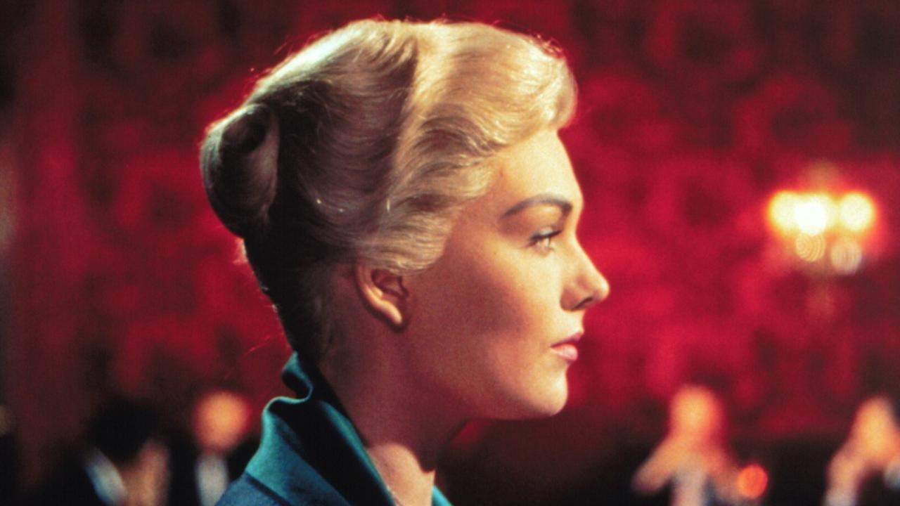 Szenenbild aus dem Film VERTIGO: Großaufnahme einer blonden Frau im rechten Vollprofil. Sie trägt die Haare hochgesteckt und ein grünes Kleid. Im Hintergrund sind unscharf Personen vor einer rottapezierten Wand und ein Leuchter zu sehen