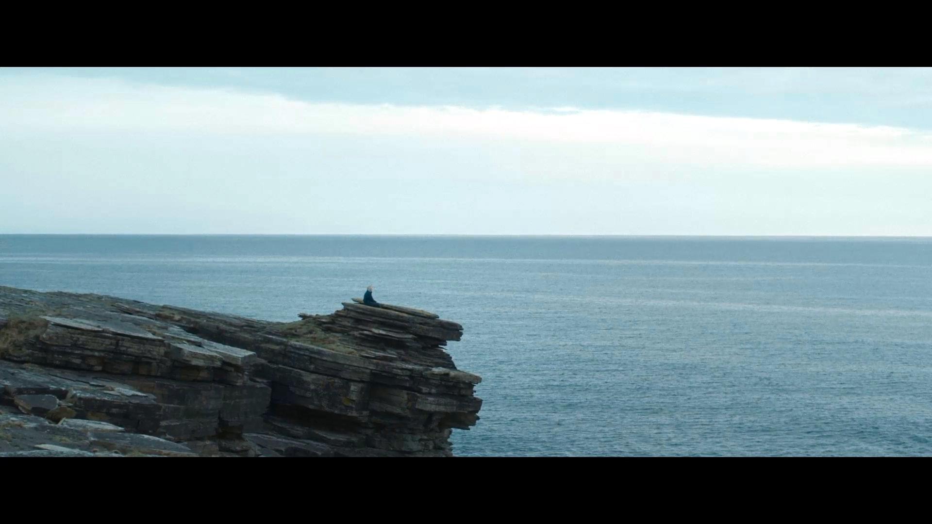 Filmstill aus dem Drama THE OUTRUN: Panoramaufnahme einer Klippe am Meer; an ihrem Rand sitzt eine menschliche Figur