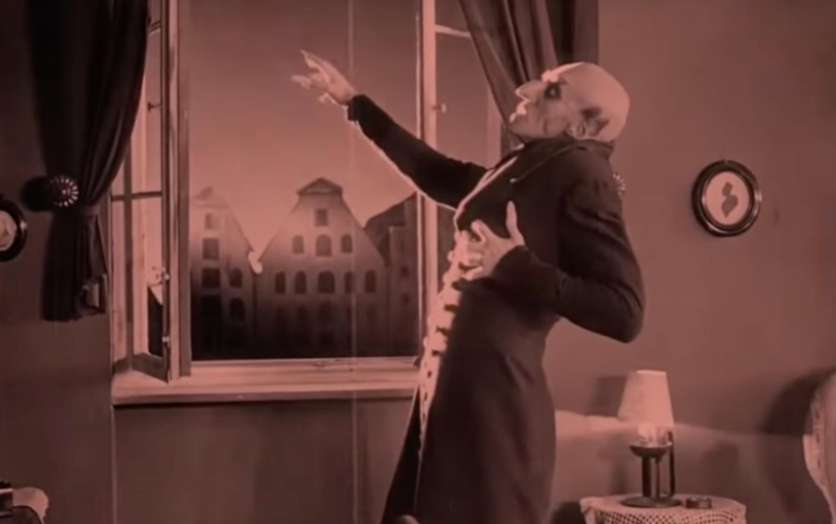 Screenshot aus dem Stummfilm NOSFERATU: Der Vampir Nosferatu steht im Halbprofil vor einem geöffneten Fenster. Er greift sich an sein Herz.