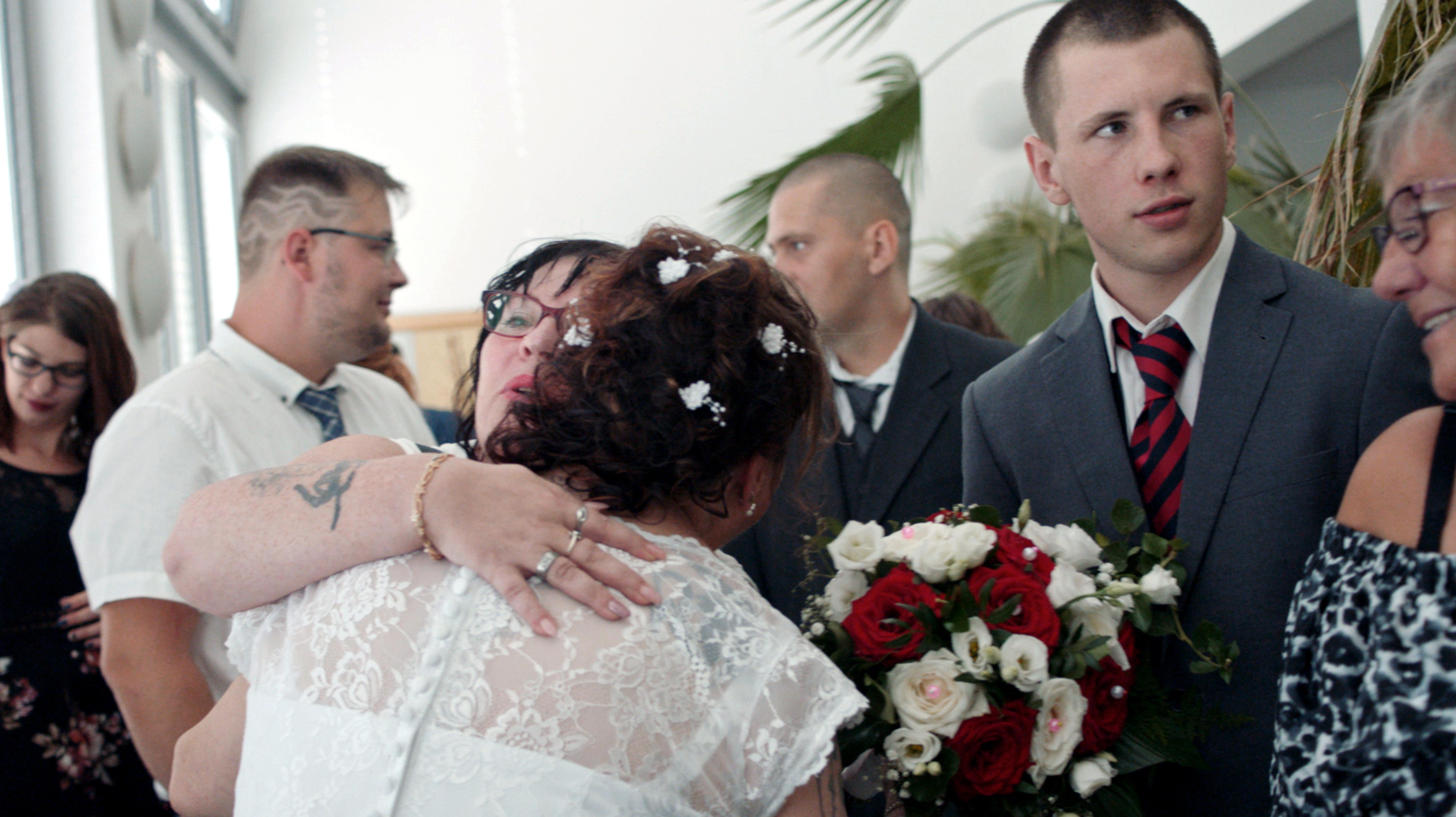 Kalle Kosmonaut, Szenenbild: Eine Frau, mit dem Rücken zum Betrachtenden, in weißem Kleid wird von einer anderen Frau umarmt. Um sie herum stehen Männer und Frauen in Festtagskleidung.