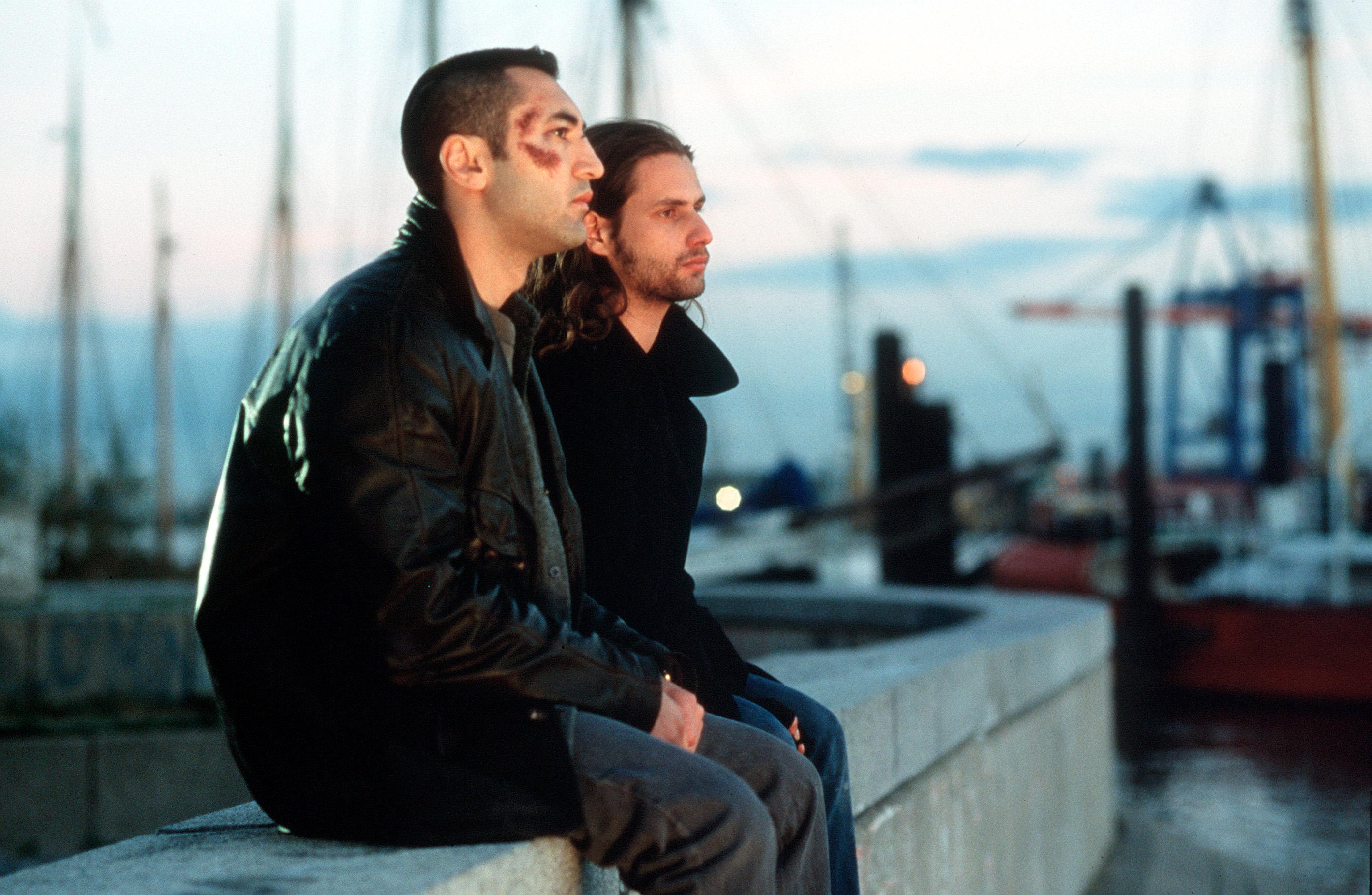 Kurz und schmerzlos, Szenenbild: Zwei junge Männer sitzen nebeneinander auf einer Mauer. Der eine Mann ist im Gesicht verletzt. Im Hintergrund sind Segelboote zu erkennen.