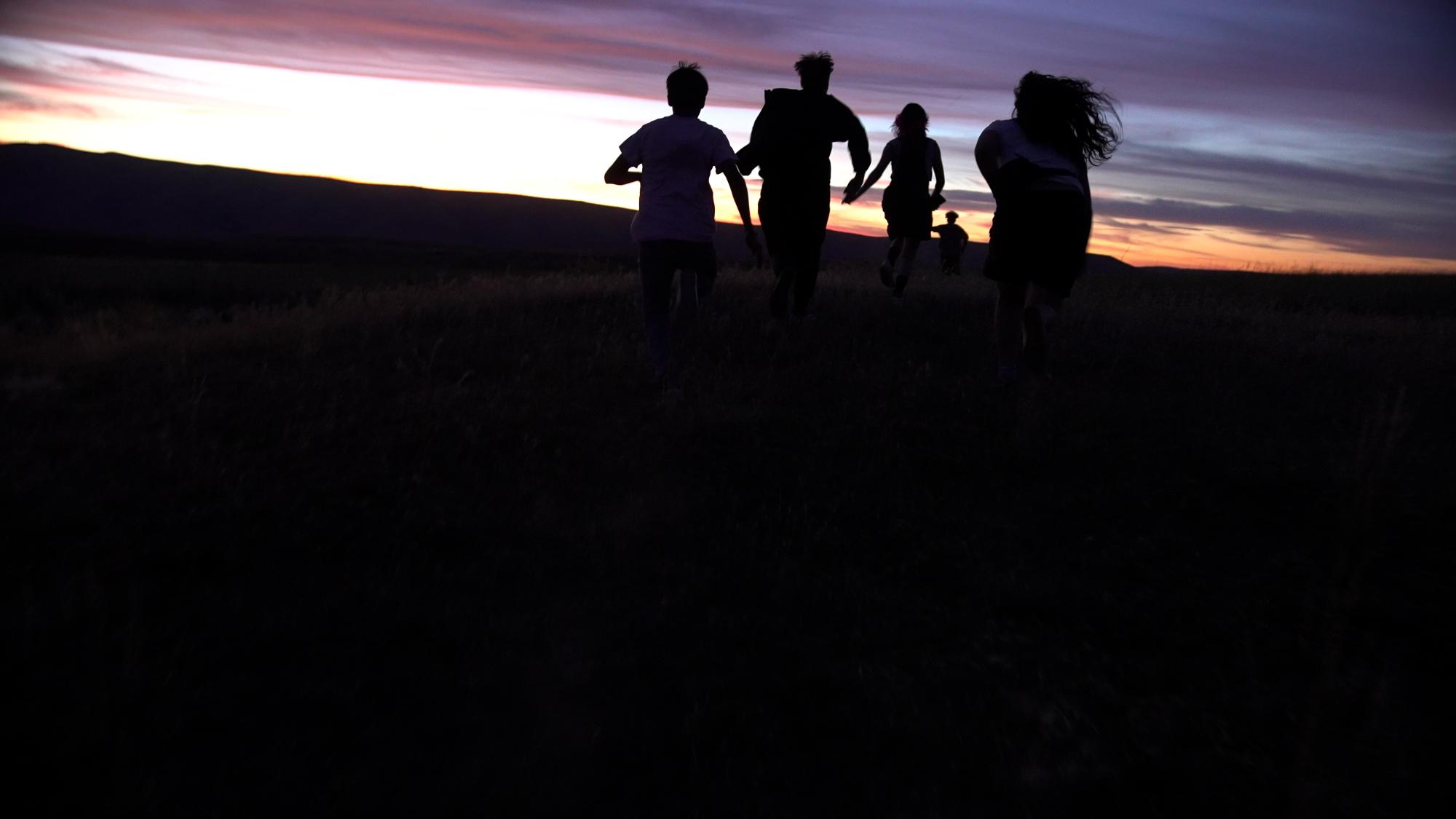 Szenenbild aus dem Roadmovie GASOLINE RAINBOW: Fünf junge Leute laufen in einer Wüstenlandschaft der untergehenden Sonne entgegen. Sie sind nur als schwarze Silhouetten erkennbar.