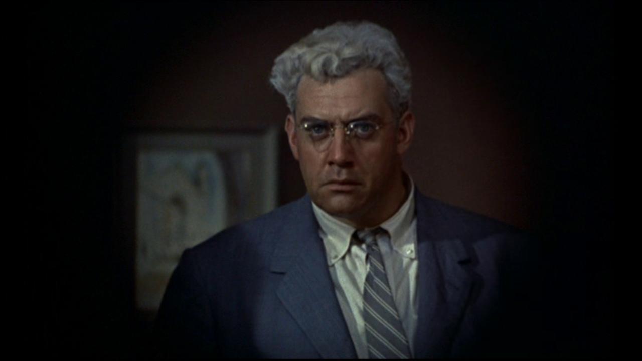 Screenshot aus dem Film "Das Fenster zum Hof": Nahaufnahme eines anzugtragenden Mannes mit grauen Haaren und Brille, der mit ernstem Gesicht aus einem dunklen Raum in Richtung Kamera schaut.