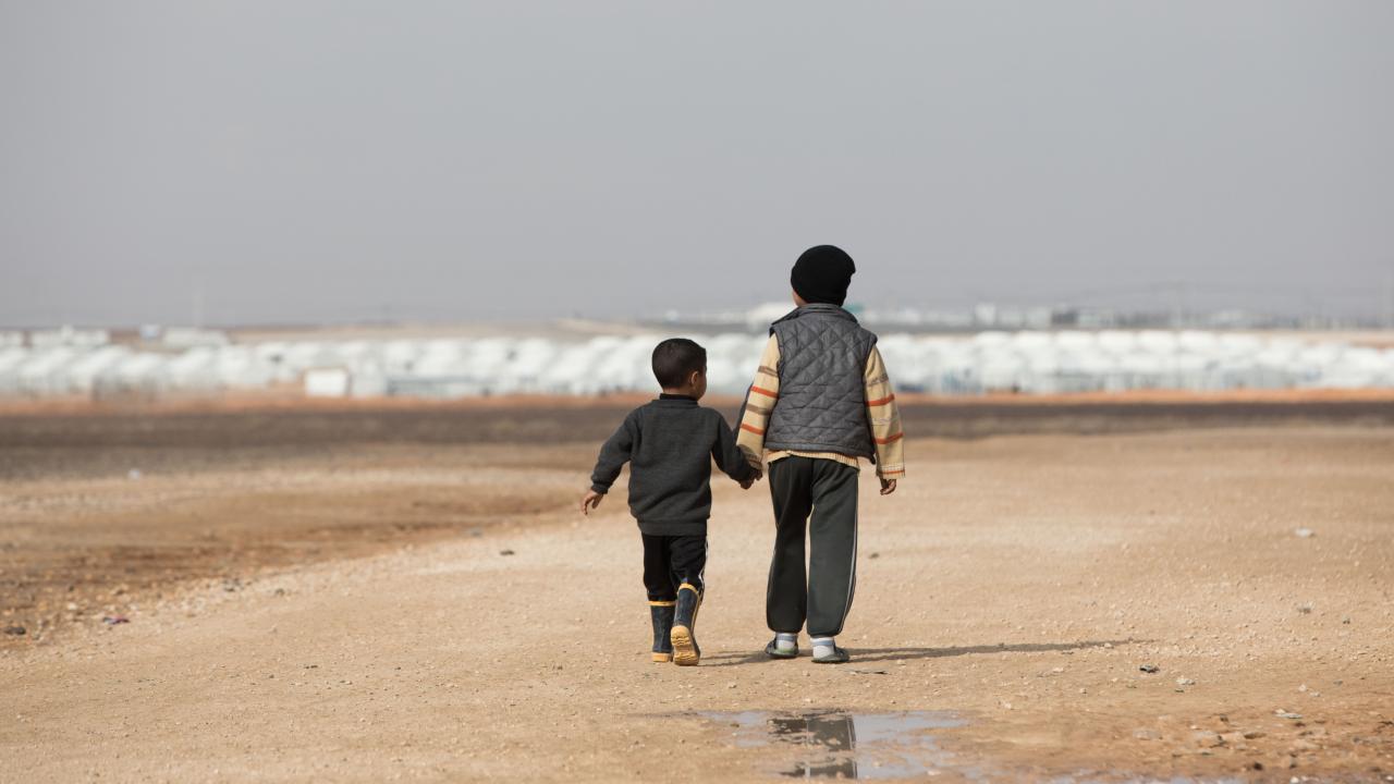 Totale: Zwei Flüchtlingskinder in der Rückenansicht laufen Hand in Hand durch eine karge, sandige Landschaft. Im Hintergrund sind schemenhaft weiße Zelte zu erkennen. Das Foto wurde am 29.01.2018 in Jordanien im Flüchtlingslager Al-Asrak aufgenommen.