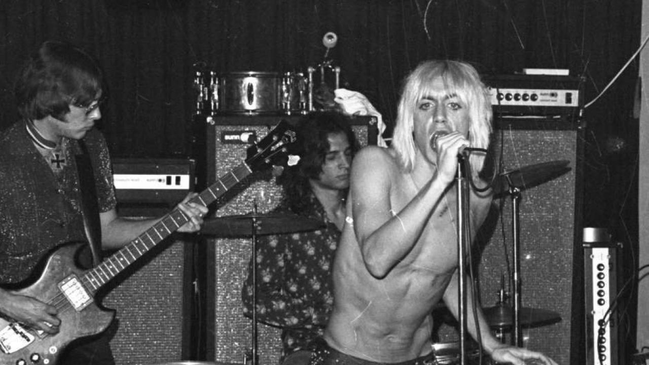 Die Band The Stooges mit Frontsinger Iggy Pop: Der Sänger steht rechts oberkörperfrei mit blonden Haaren und Mikrofone in der Hand und vor dem Gesicht. Der Bass-Spieler steht links und guckt konzentriert auf seiner Guitarre runter. Hinter sitzt der Schlagzeugspieler hinter seinem Instrument. Im Hintergrund sind große Lautsprecheranlagen. 