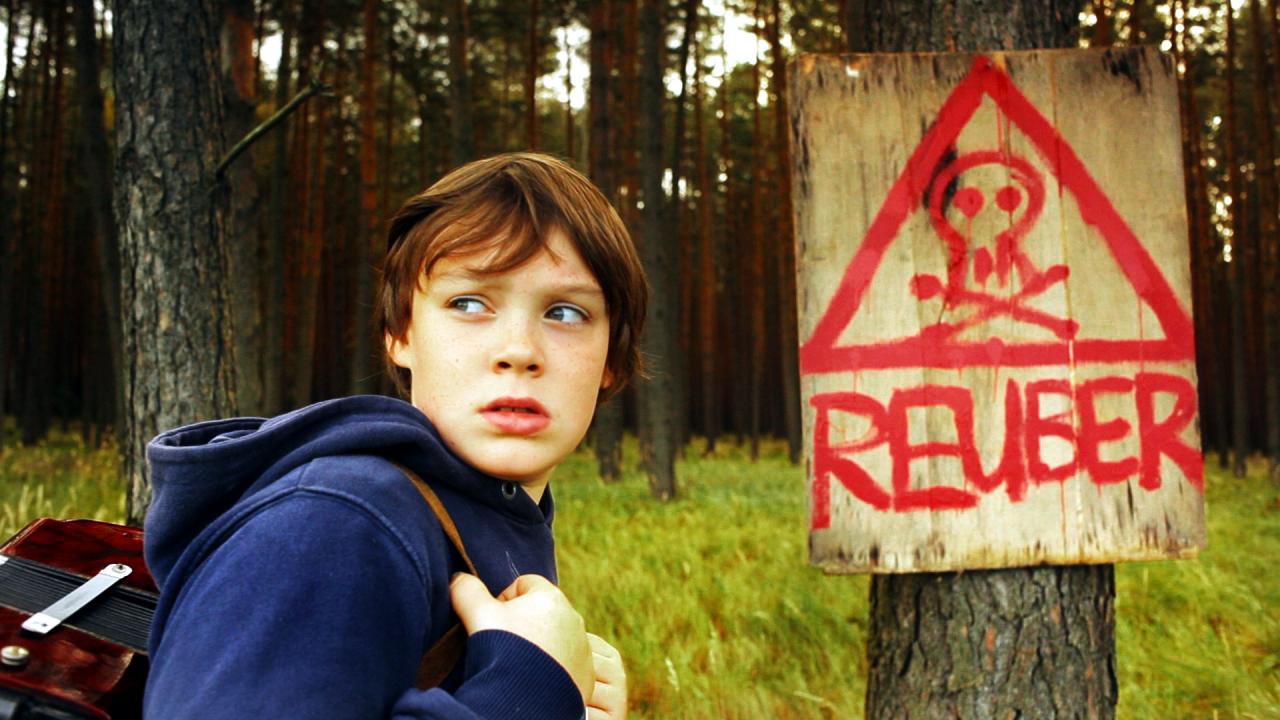 Szenenbild aus dem Spielfilm "Reuber": Ein Junge im blauen Hoodie steht in einem Wald. Er wirkt ängstlich und schaut nach hinten. Rechts von ihm hängt an einem Baum ein Schild, auf dem mit roter Farbe ein Totenkopf und das Wort "Reuber" gemalt ist.