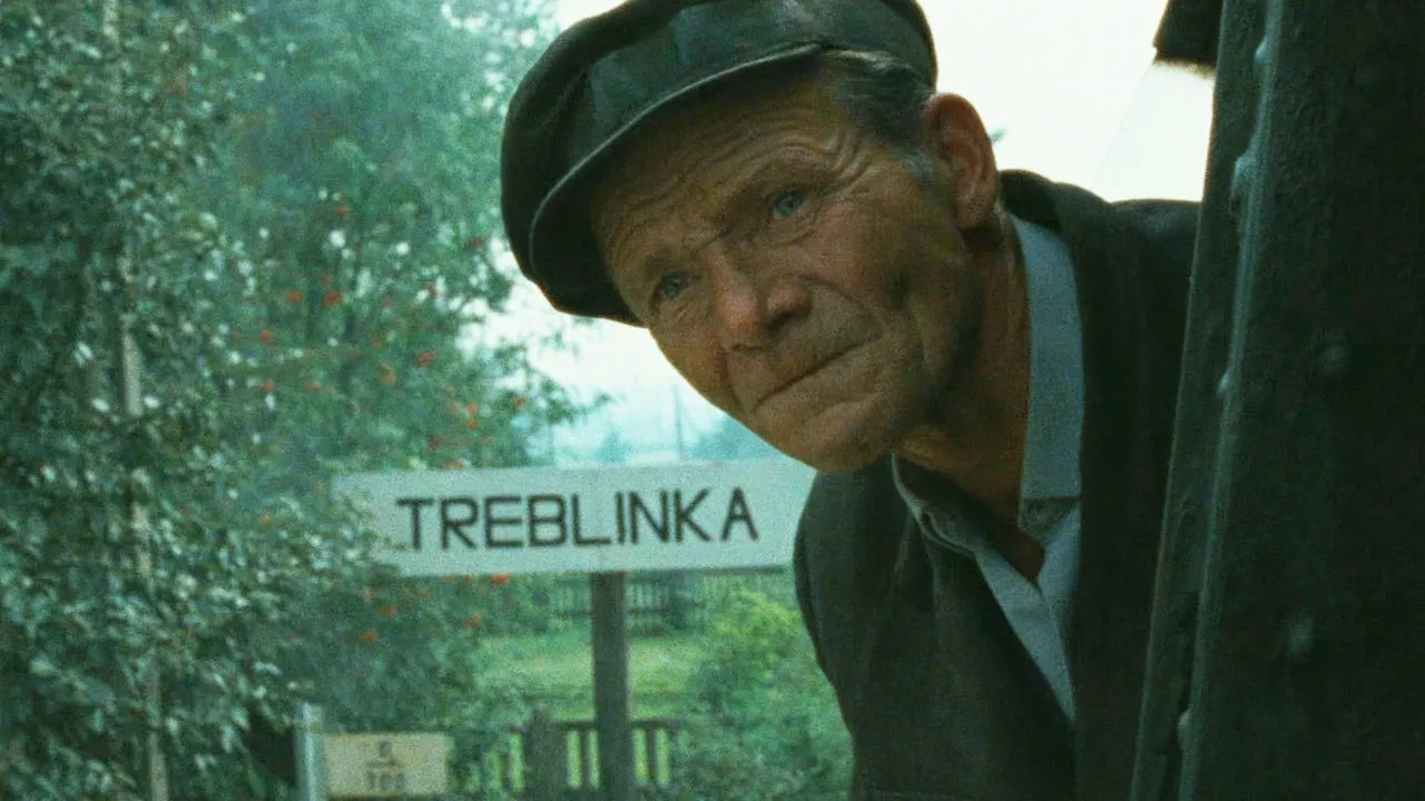 Ein alter Mann mit Mutze schaut aus einer Lok direkt in die Kamera. Unten links ist ein Schild mit dem Schriftzug "Treblinka". 