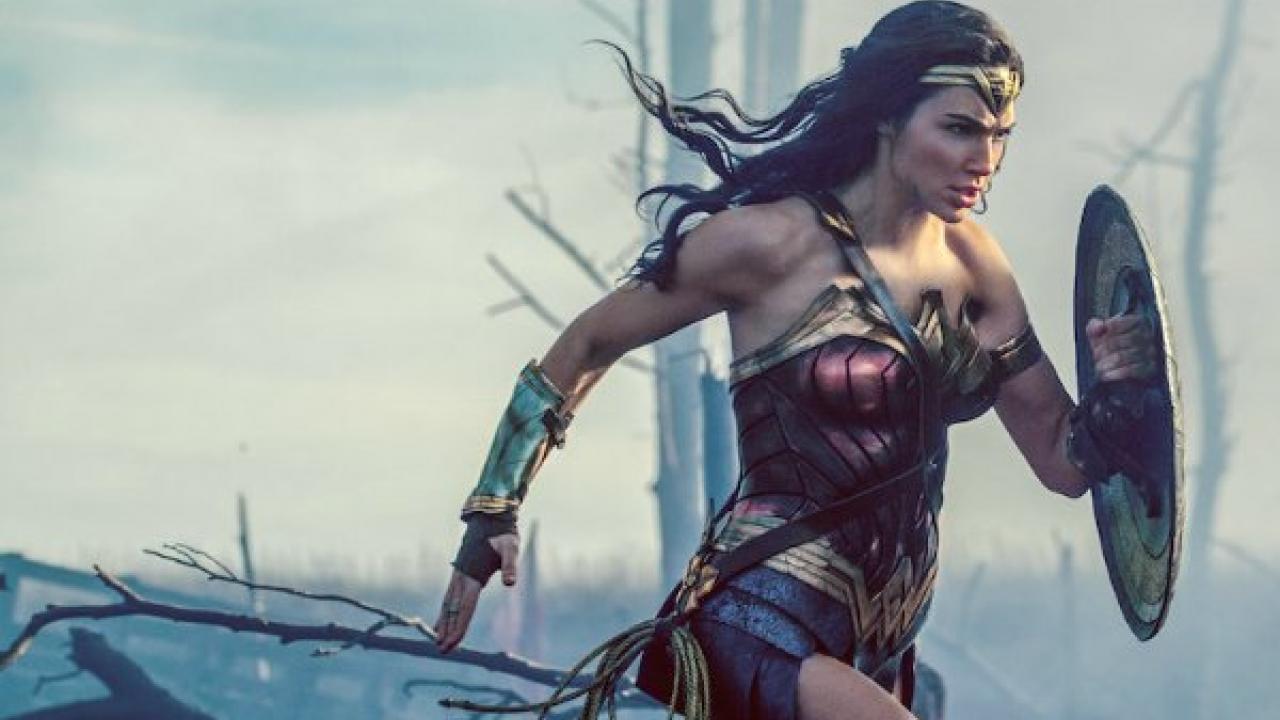 Wonderwoman in rot-goldener Rüstung rennt mit ihrem Schild vor ihr und ihren Lasso an der Hüfte geschnallt. Sie guckt konzentriert. Im Hintergrund ist eine graue leere Kreigslandschaft. 