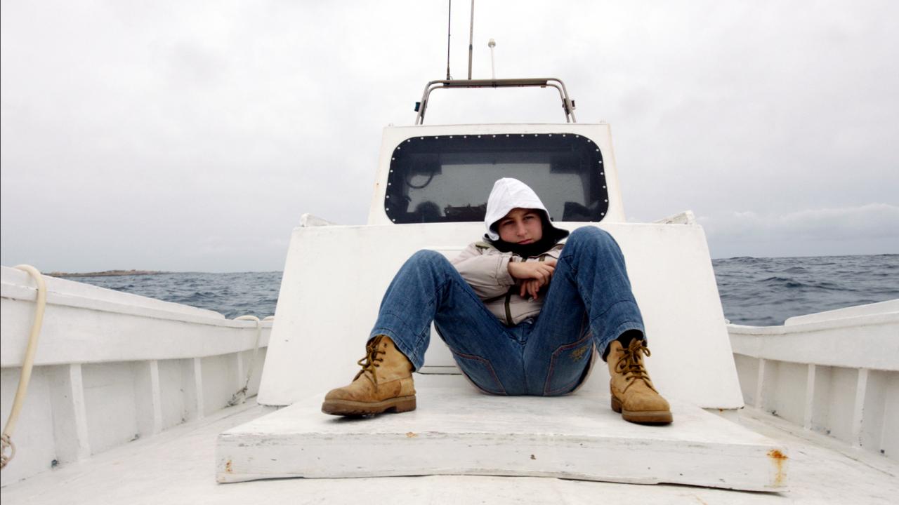 Szenenbild aus dem Dokumentarfilm "Seefeuer": Ein Junge im Teenageralter sitzt breitbeinig auf einem Boot, hinter ihm ist die Steuerkabine zu sehen und das offene Meer.