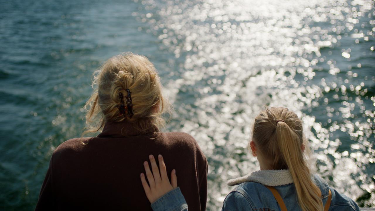 Rückenansicht eines blonden Mädchens und einer älteren Frau, die gemeinsam auf das Meer schauen. Das Kind hat ihre Hand auf den Rücken der Frau gelegt.