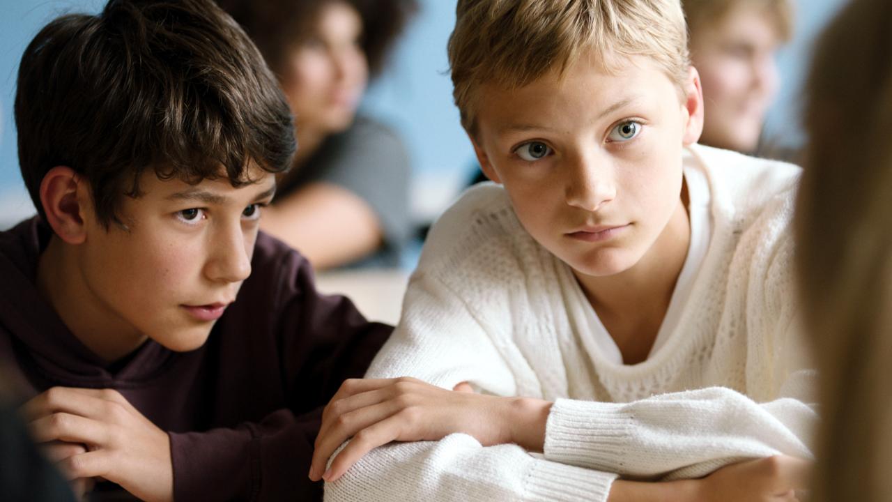 Szenenbild aus dem Film "Close": Nahaufnahme: Zwei Jungen im Teenageralter – der eine dunkelhaarig, der andere blond – sitzen in einem Klassenraum nebeneinander und hören jemanden zu. 