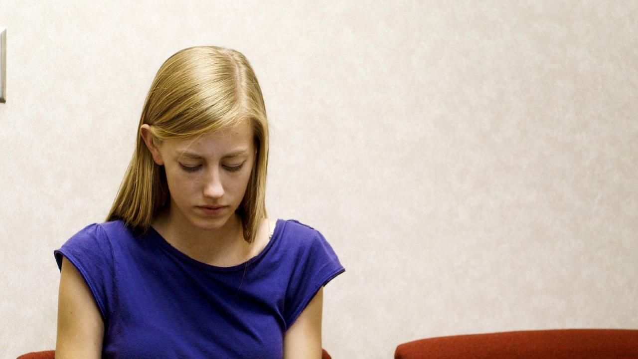 Ein Mädchen mit blonden Haaren sitzt in einem Wartezimmer und schaut nach unten. Sie trägt ein blaues T-Shirt.