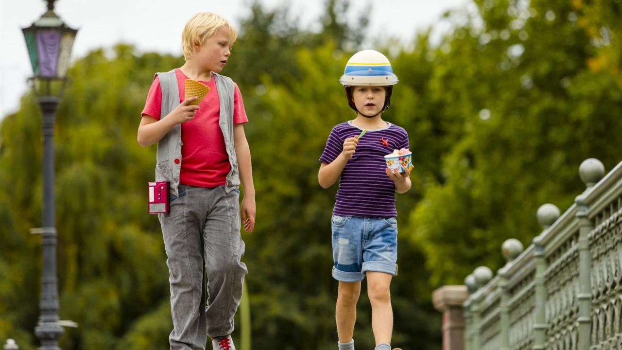 Zwei Jungen gehen auf einer Brücke, einer isst ein Eis, der andere trägt einen Helm und hält einen Becher. Grüne Bäume im Hintergrund.