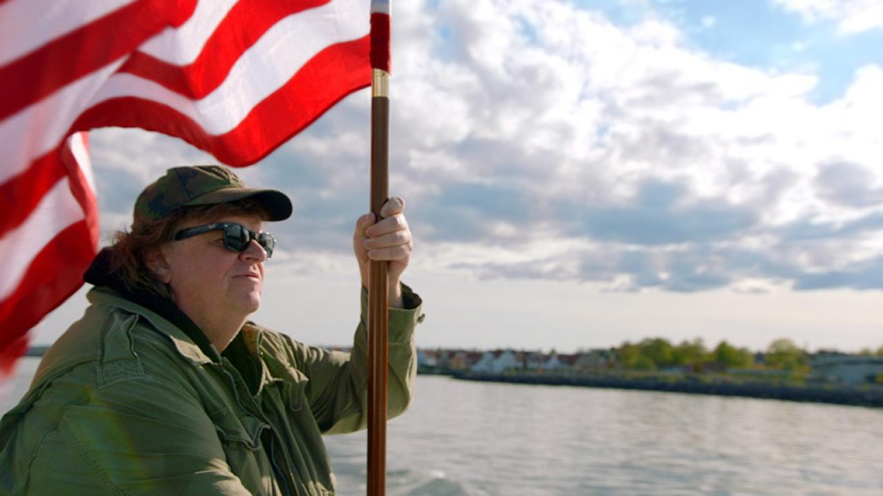 Michael Moor trägt die amrikanischen Flagge und steht auf einem Boot. 