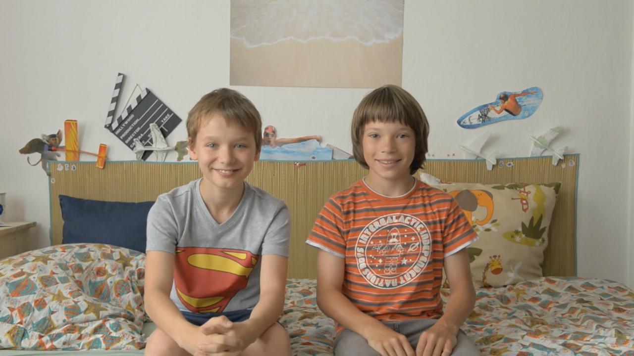 Zwei lachende Jungen sitzen auf einem Bett in einem farbenfrohen Kinderzimmer. Spielzeug und Poster im Hintergrund.