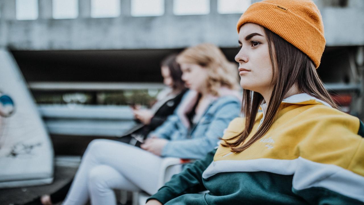 Junge Frau mit gelber Mütze und Hoodie sitzt draußen, während zwei Freundinnen im Hintergrund auf ihre Handys schauen.