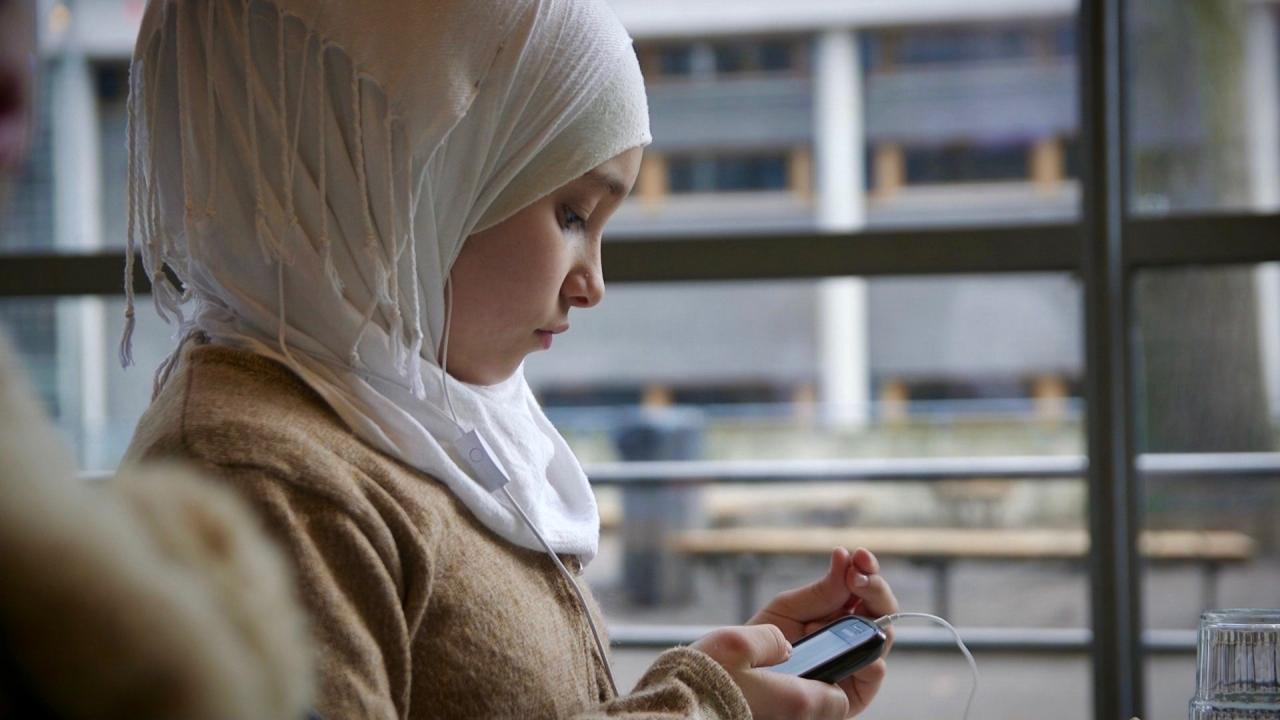 Mädchen mit Kopftuch sitzt am Fenster und benutzt ein Smartphone. Glas Wasser im Vordergrund.