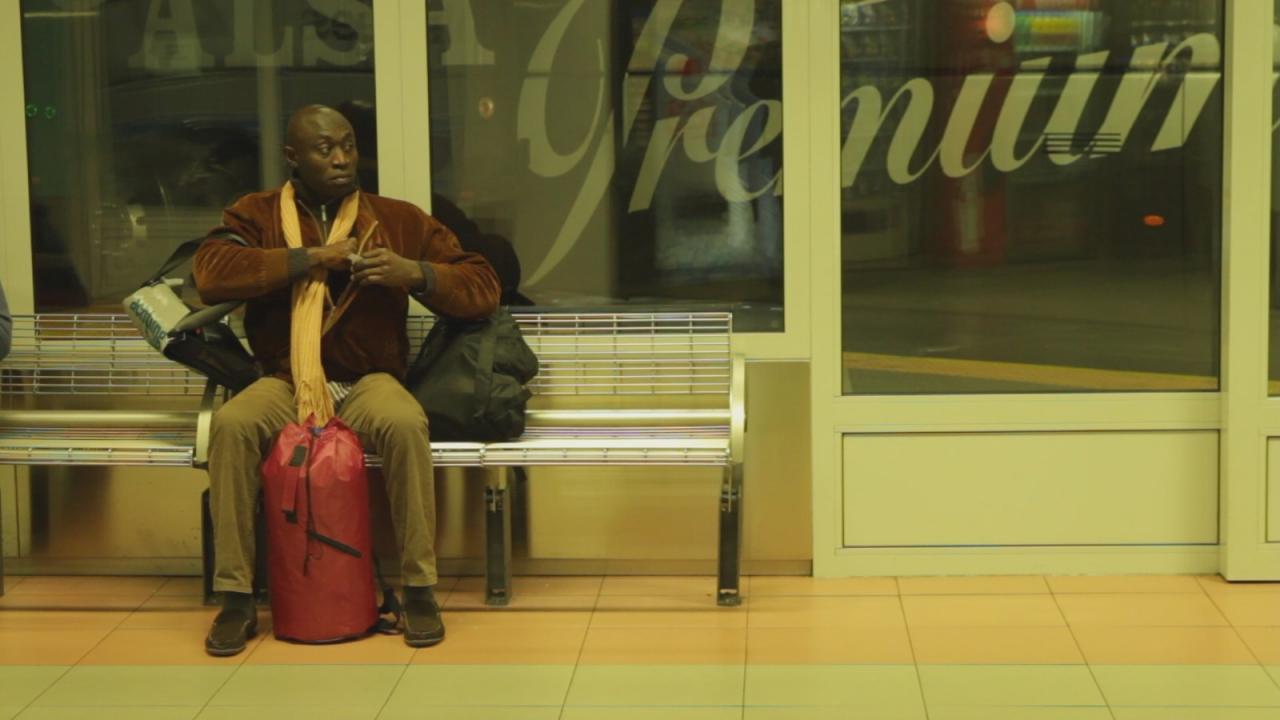 Mann mit brauner Jacke und gelbem Schal sitzt auf einer Bank in einer Wartehalle mit roter Tasche und Rucksack.