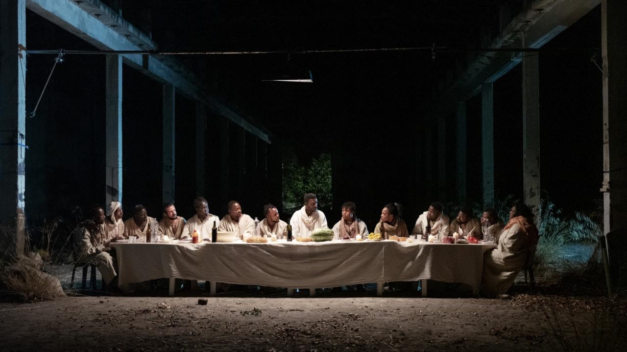 Gruppe von Menschen sitzt an einem langen Tisch in einer dunklen, verlassenen Halle, Kerzen und Essen auf dem Tisch. Nachstellung des "letzten Abendmahls"