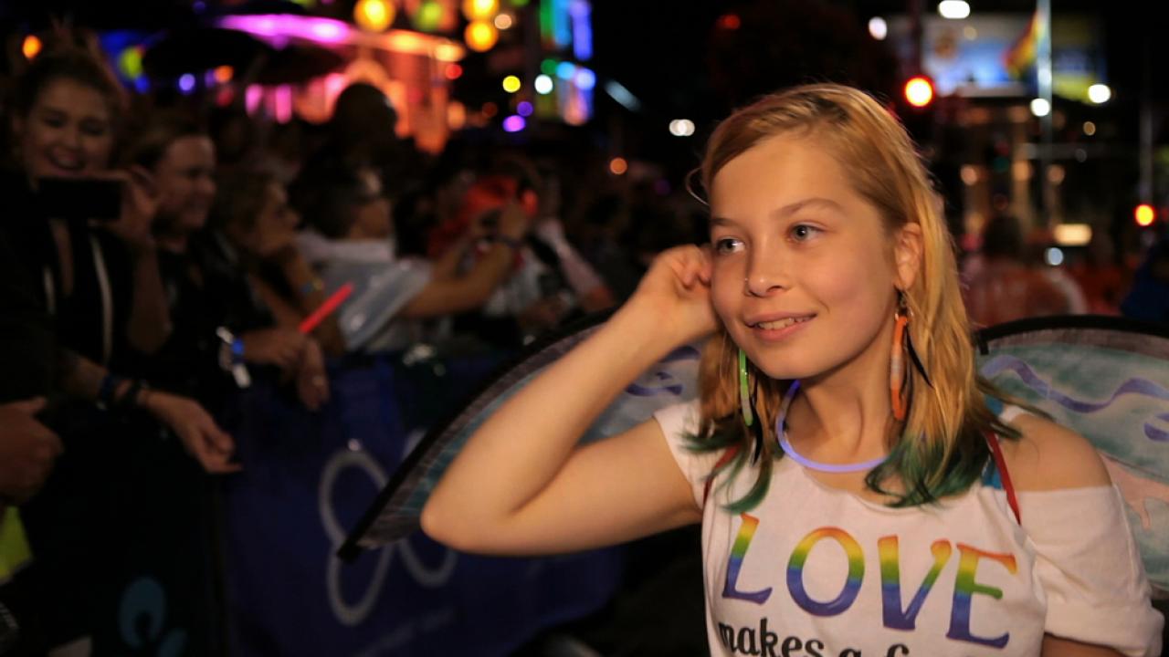 Mädchen mit regenbogenfarbenem T-Shirt bei Nachtparade, lächelt und trägt bunte Accessoires. Zuschauer im Hintergrund.