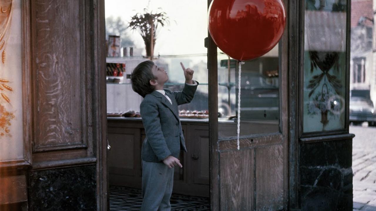 Ein kleiner Junge im grauen Anzu steht in der Tür eines Cafés. Mit erhobenen Finger scheint er zu einem roten Ballon zu sprechen.