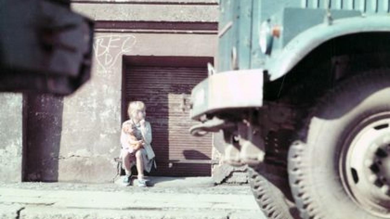 Halbtotale: Ein kleines Mädchen sitzt allein an einer Hauswand. Rechts sieht man die Reifen und die Motorhaube eines Lastwagens.