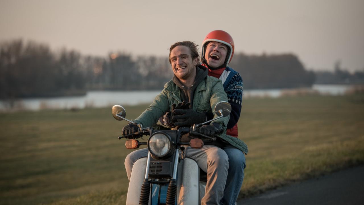 Zwei lachende Männer fahren auf einem Motorrad, der Beifahrer trägt einen roten Helm, Landschaft im Hintergrund.