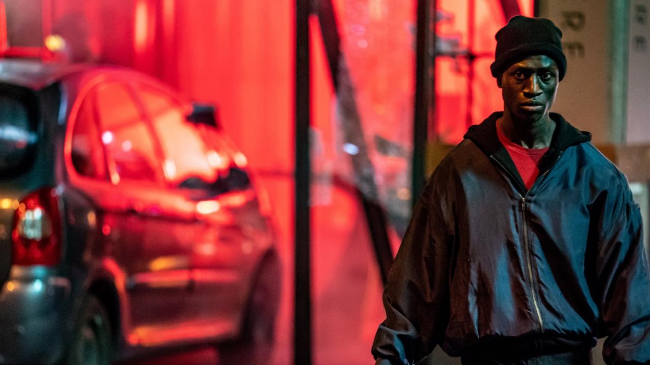 Szenenbild aus dem Drama BERLIN ALEXANDERPLATZ: Ein junger Mann of Colour läuft direkt auf die Kamera zu. Er hält eine Clownsmaske in der Hand. Im Hintergrund steht ein Auto mit roten Lichter belichtet. 