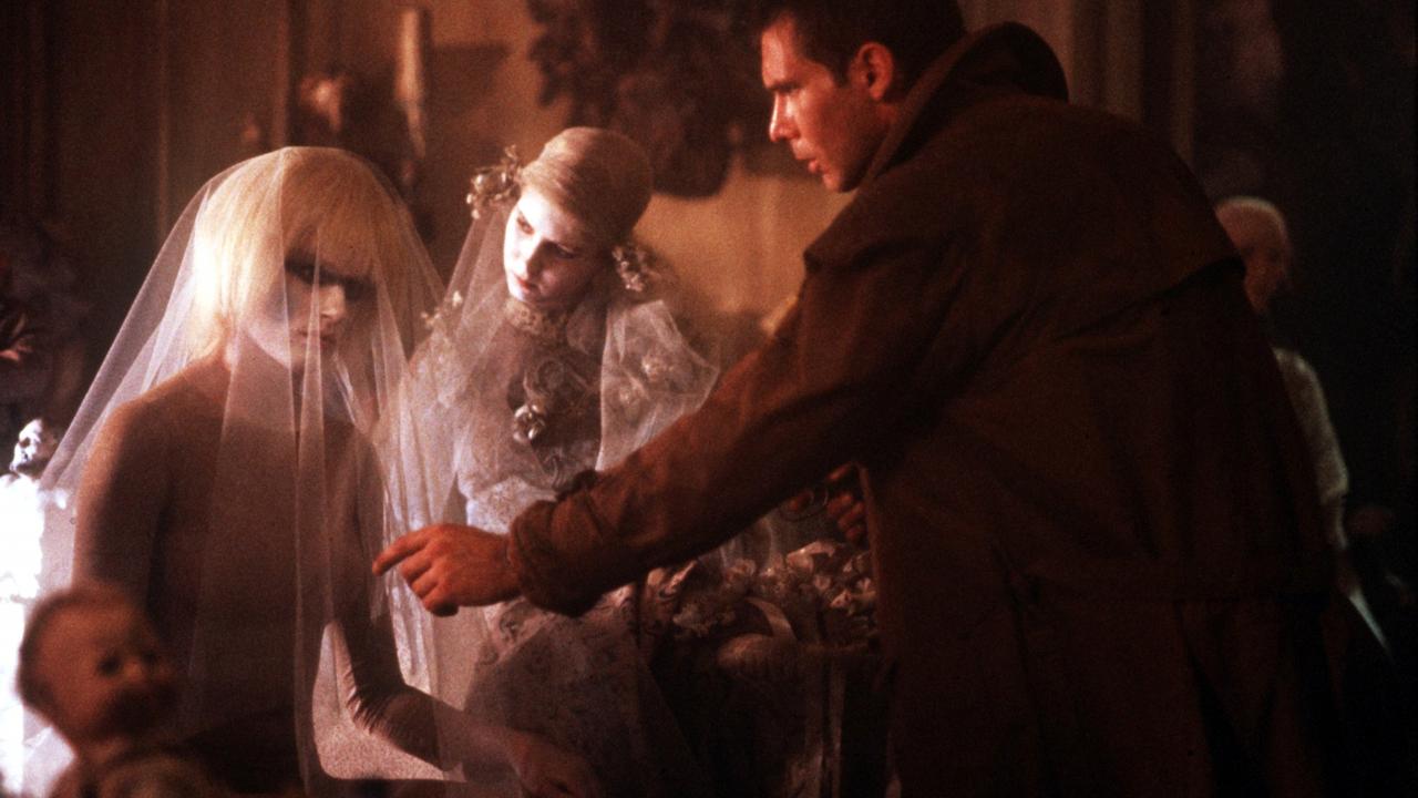 Szenenbild aus dem Science-Fiction-Film BLADE RUNNER: Ein Mann in einem langen Mantel streckt die Hand aus in Richtung einer Frau mit blondem Haar, die einen durchsichtigen Schleier trägt. Im Hintergrund steht eine weitere Frau in einem weißen, kunstvollen Kleid.