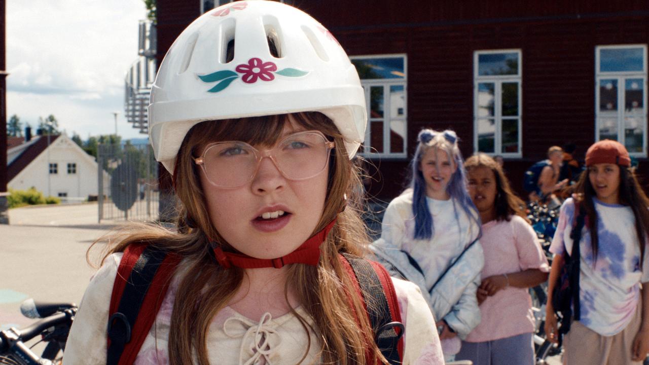 Szenenbild aus dem Spielfilm DANCING QUEEN: Nahaufnahme eines jungen Mädchens mit Brille und weißem Fahrradhelm. Sie steht auf einem Schulhof. Im Hintergrund sind vier weitere Teenagerinnen zu sehen.