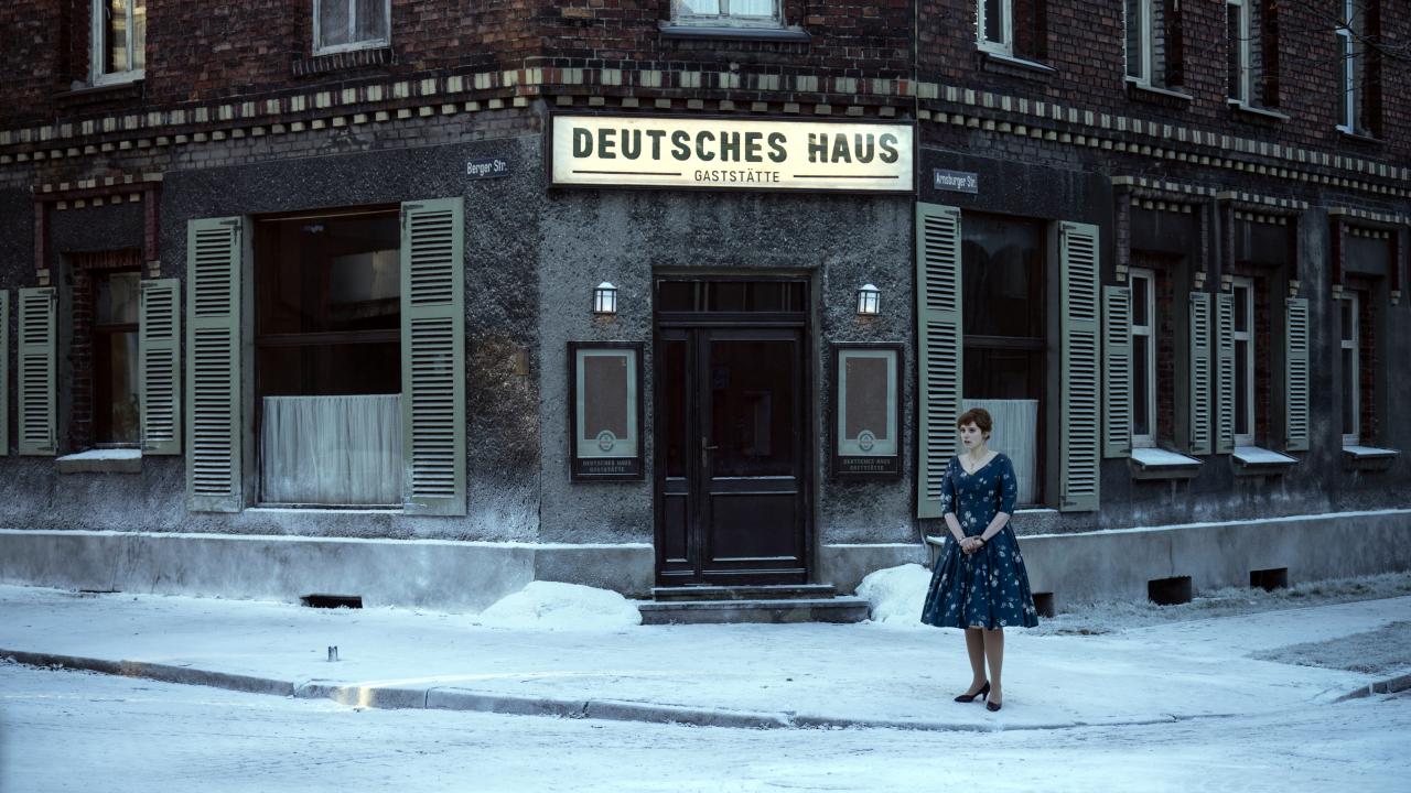 Szenenbild aus der Serie DEUTSCHES HAUS: Eine junge Frau, gekleidet im Stil der 1960er-Jahre, steht im Winter ohne Mantel vor einer Gaststätte, die "Deutsches Haus" heißt.