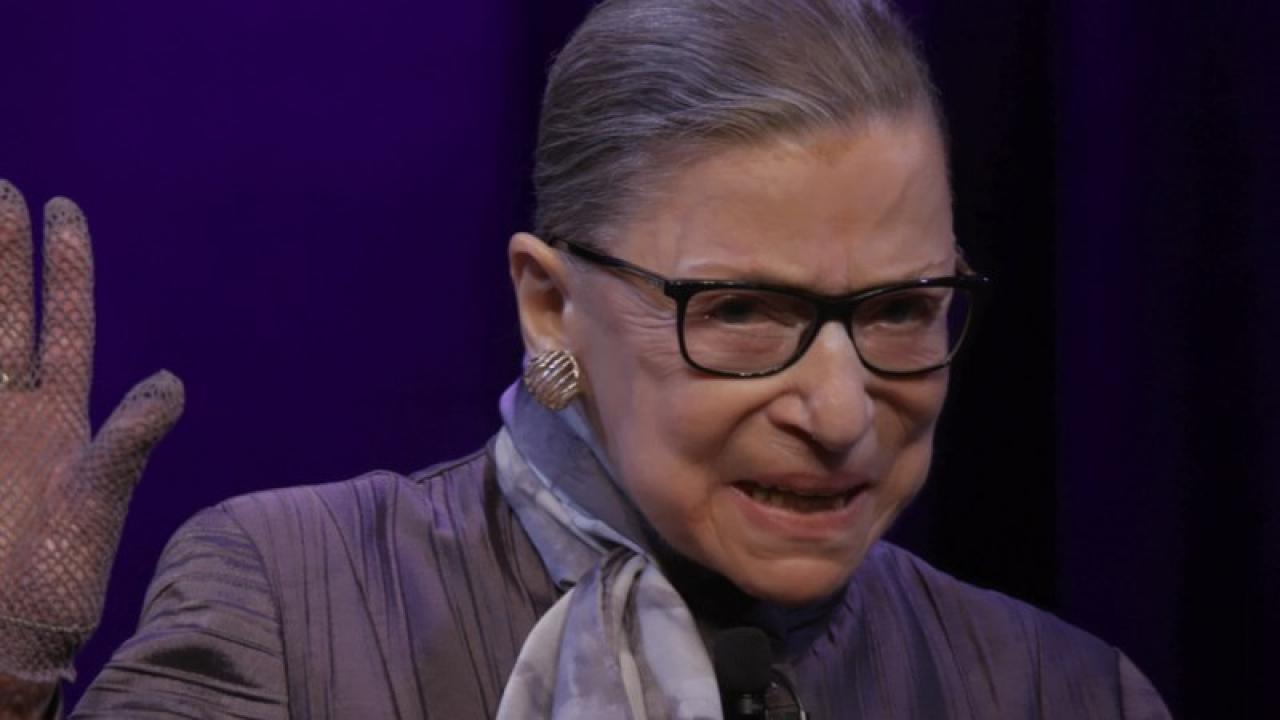 Porträt von Ruth Bader Ginsburg. Eine Dame um die 80 mit grauen zückgekämmten Haaren und schwarzer Brille, winkt lächelnd.