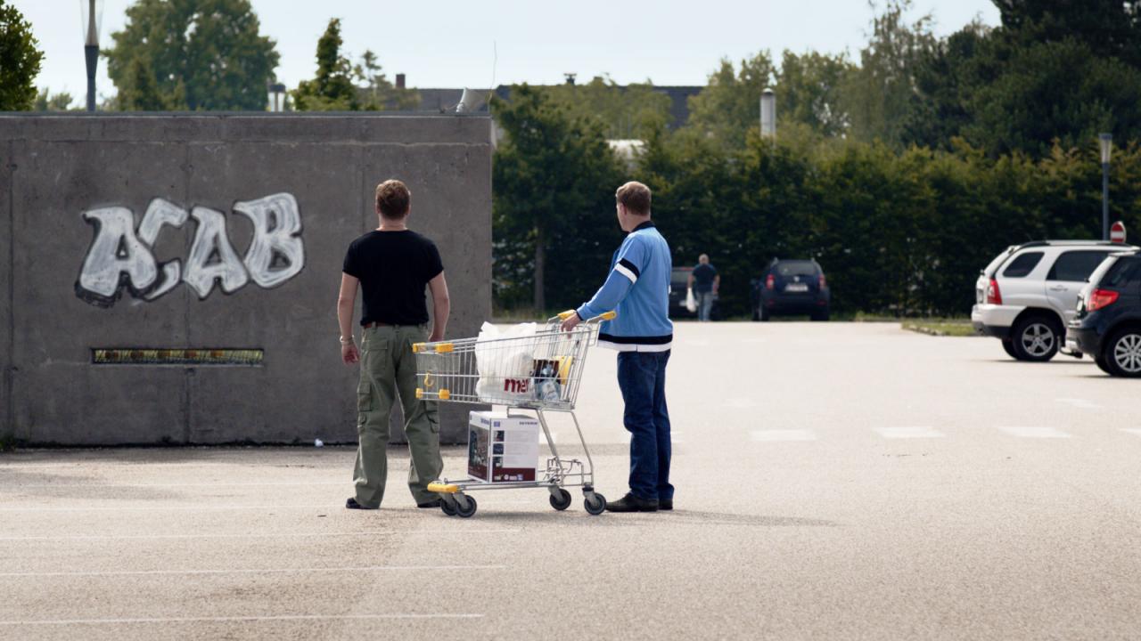 Einer von uns (Szenenbild): Zwei Männer stehen auf einen Parkplatz, einer schiebt ein Einkaufswagen. Mit den Rücken zu uns schauen sie auf einer Wand wo es groß "ACAB" geschrieben wurde. 