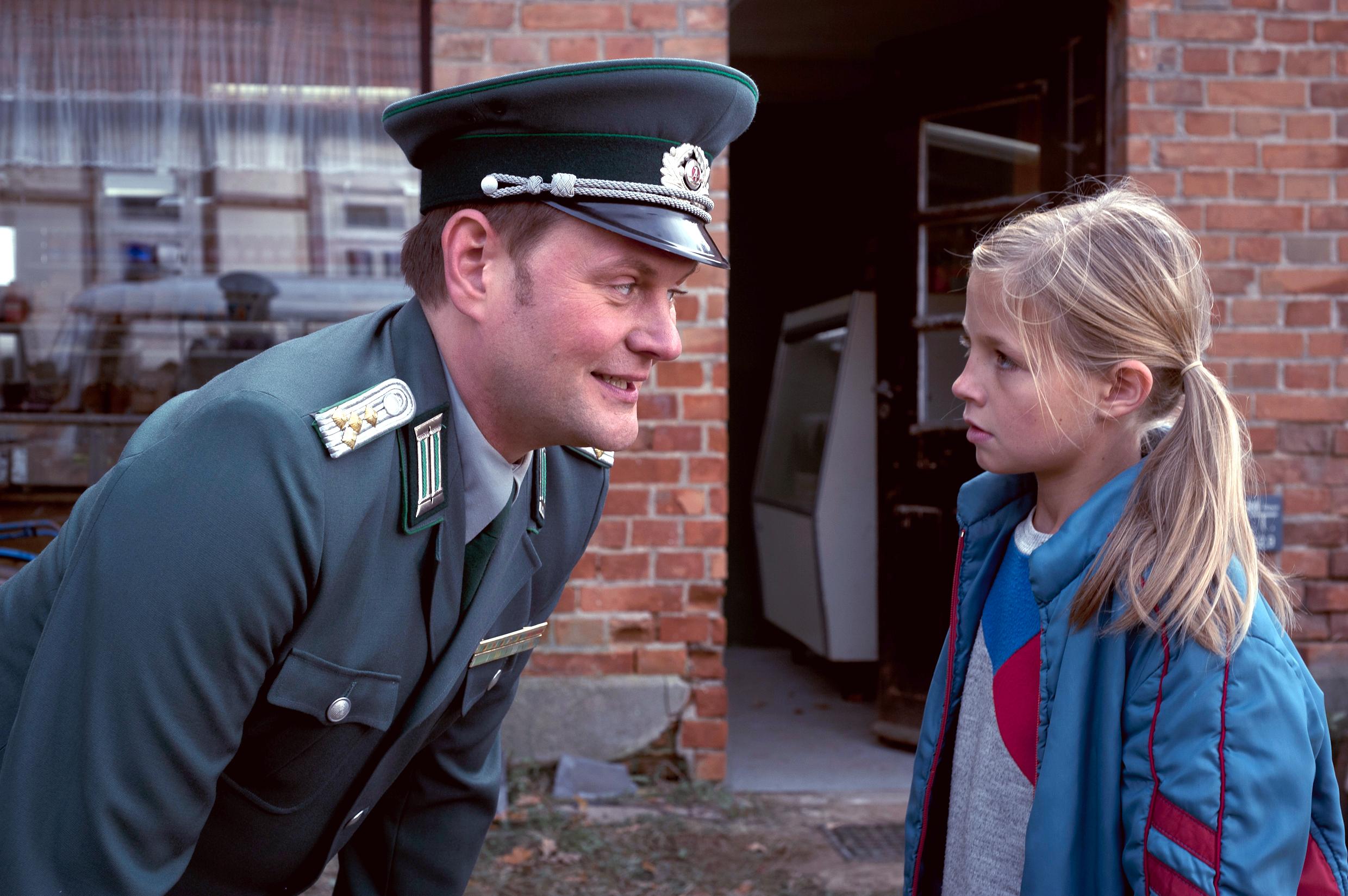 Szenenbild aus dem Kinderfilm "Sputnik": Ein DDR-Volkspolizist beugt sich zu einem Mädchen mit blonden Zöpfen herunter und spricht mit ihm.