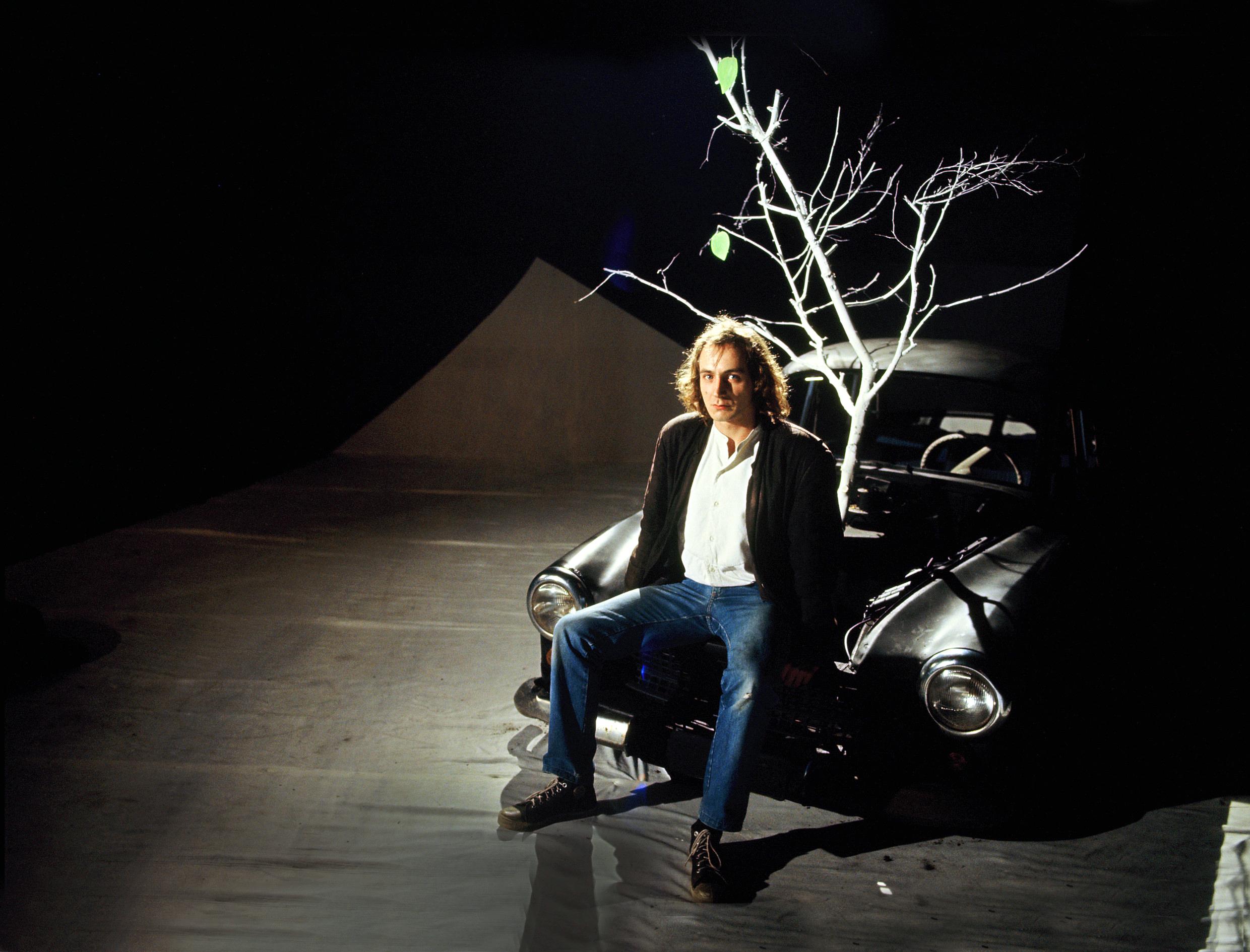 Szenenbild aus dem Spielfilm "Stilles Land": Eine Theaterbühne mit dunklem Hintergrund. Ein Mann in Jeans, weißem Hemd und dunkler Jacke sitzt auf der Motorhaube eines Autos, ein kahler Baum im Hintergrund.